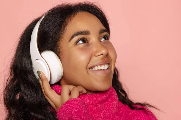 Close-up young pretty dark skinned smiling girl in warm knitted sweater and headphones isolated on pink background. — Zdjęcie stockowe