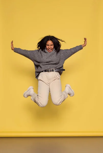 Retrato de larga duración de la joven chica de piel oscura feliz en jersey de punto cálido saltando aislado sobre fondo amarillo. — Foto de Stock
