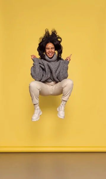 Retrato de larga duración de la joven chica de piel oscura feliz en jersey de punto cálido saltando aislado sobre fondo amarillo. — Foto de Stock