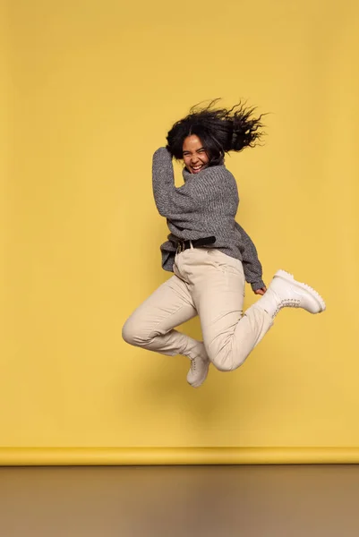Full-length portrait of young happy dark skinned girl in warm knitted sweater jumping isolated on yellow background. — Fotografia de Stock