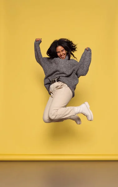 Full-length portrait of young happy dark skinned girl in warm knitted sweater jumping isolated on yellow background. — Fotografia de Stock