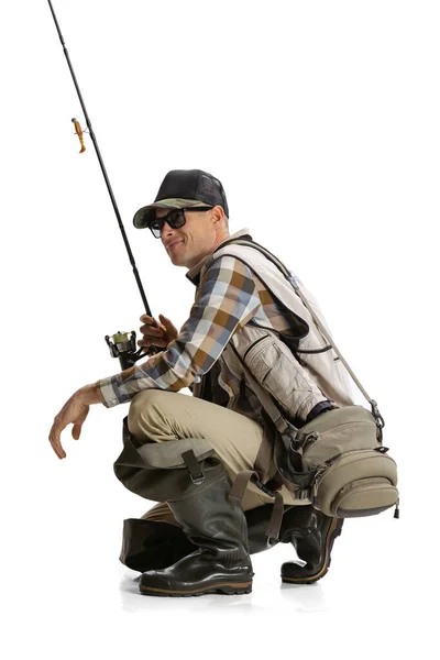 Portrait of young man, professional fisherman with fishing rod, spinning and equipment sitting isolated over white studio background — Stock Photo, Image