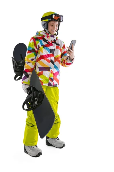 Menina feliz em sportswear brilhante, óculos e capacete usando telefone com snowboard isolado no fundo do estúdio branco. Conceito de esportes de inverno — Fotografia de Stock