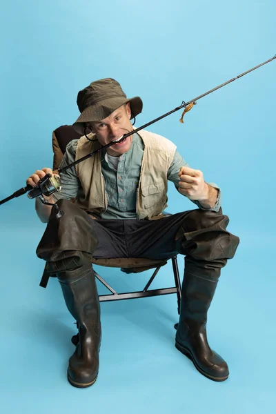 Retrato de joven, pescador con caña de pescar, spinning y equipo sentado aislado sobre fondo azul estudio — Foto de Stock