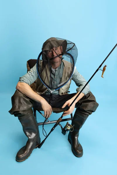 Retrato de joven, pescador con caña de pescar, spinning y equipo sentado aislado sobre fondo azul estudio —  Fotos de Stock