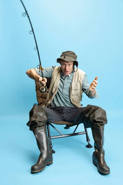 Surprised, shocked young man, fisherman with fishing rod, spinning and equipment sitting isolated over blue studio background — Stockfoto