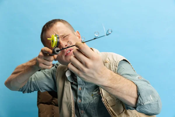 Funny young man, fisherman with fishing accessories sitting on chair isolated over blue studio background — стоковое фото