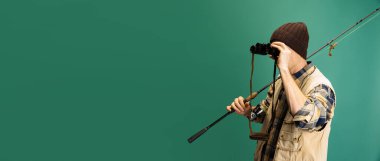 Genç adamın stüdyo fotoğrafı, balıkçı aksesuarları, yeşil arka planda izole edilmiş spor kıyafetleri giyen balıkçı.