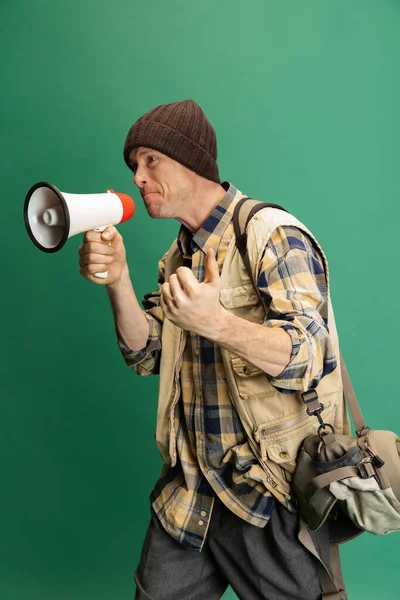 Close-up of young man, fisherman with fishing accessories wearing sport style clothes shouting at megaphone isolated over green background — Stockfoto