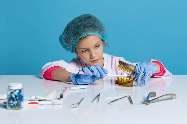 Emotional cute little girl, child in image of dentist doctor wearing white lab coat and gloves isolated on blue studio background — Fotografia de Stock