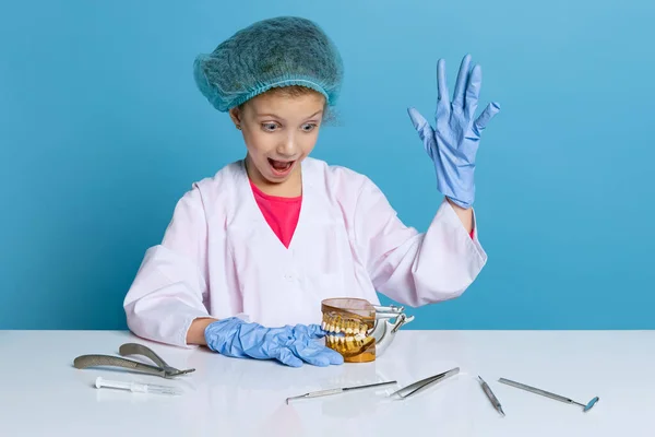 感情上可爱的小女孩，身穿白色实验室外套，戴着手套的牙医形象中的孩子被隔离在蓝色工作室的背景下 — 图库照片
