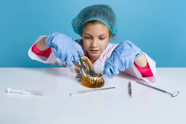 Emotional cute little girl, child in image of dentist doctor wearing white lab coat and gloves isolated on blue studio background — Zdjęcie stockowe