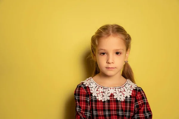 Полудлинный портрет милой маленькой девочки, ученица в клетчатом платье позирует изолированно на желтом фоне. Концепция детства, эмоции, учеба — стоковое фото