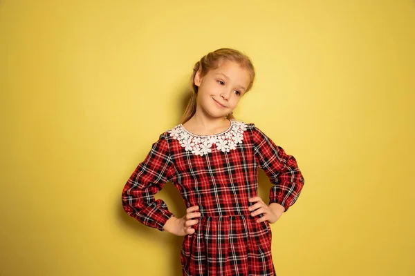 Полудлинный портрет милой маленькой девочки, ученица в клетчатом платье позирует изолированно на желтом фоне. Концепция детства, эмоции, учеба — стоковое фото