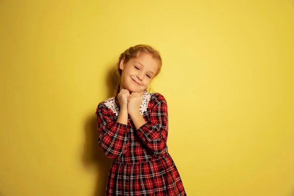 Retrato de media longitud de una linda niña, alumna con vestido a cuadros posando aislada sobre fondo amarillo. Concepto de infancia, emociones, estudio — Foto de Stock