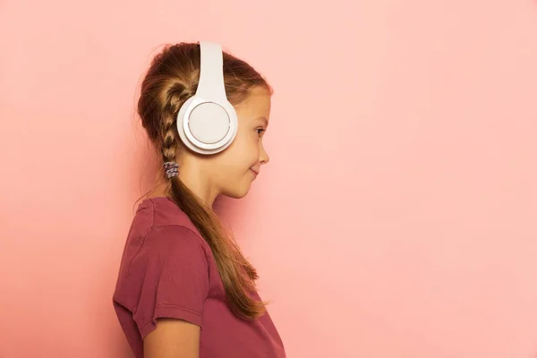 Profile view of little girl wearing t-shirt listening to music in headphones isolated on pink background. Concept of childhood, emotions, study — Stock Photo, Image