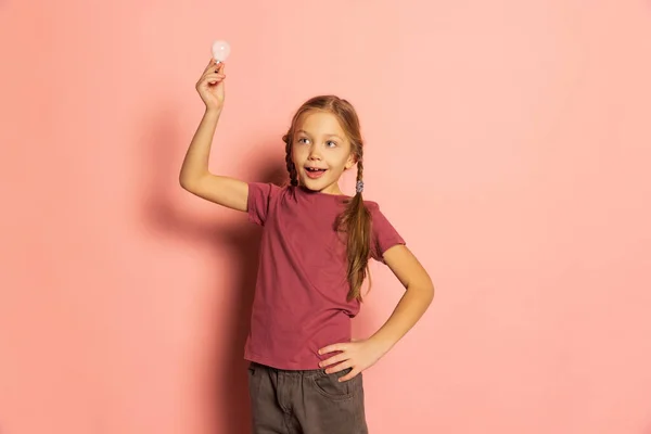 Емоційна мила дівчинка, зіниця тримає лампочку, позує ізольовано на рожевому фоні. Концепція дитинства, емоцій, навчання — стокове фото