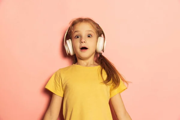 Zaskoczona dziewczynka w t-shircie słuchająca muzyki w słuchawkach odizolowanych na różowym tle. Koncepcja dzieciństwa, emocje, nauka — Zdjęcie stockowe