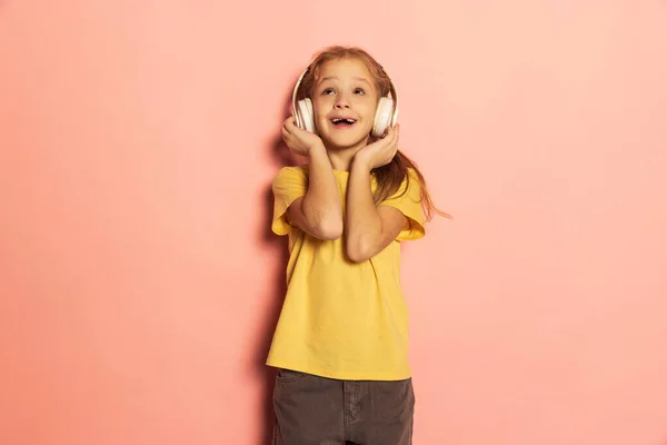 Happy little girl wearing t-shirt listening to music in headphones isolated on pink background. Concept of childhood, emotions, study — Stock Photo, Image
