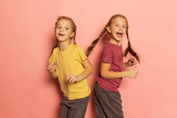 Веселі маленькі дівчата, сестри або брати і сестри в повсякденному одязі, танцюють ізольовано на рожевому тлі. Поняття дитинства, емоції — стокове фото