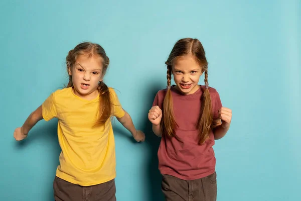 Дві роздратовані маленькі дівчата, сестри або брати і сестри носять одяг в повсякденному стилі ізольовані на синьому фоні. Поняття дитинства, емоції — стокове фото