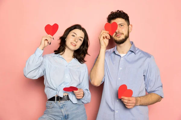 Fiatal és boldog férfi és nő kezében üdvözlőkártyák alakú szív elszigetelt rózsaszín trendi színes háttér. Érzelmek, ifjúság, szerelem és életmód koncepció — Stock Fotó