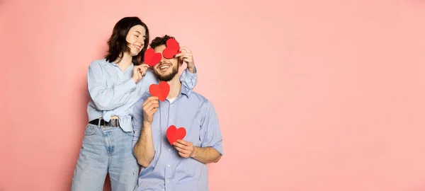 Szórólap. Fiatal és boldog férfi és nő kezében üdvözlőkártyák alakú szív elszigetelt rózsaszín trendi színes háttér. Érzelmek, ifjúság, szerelem és életmód koncepció — Stock Fotó