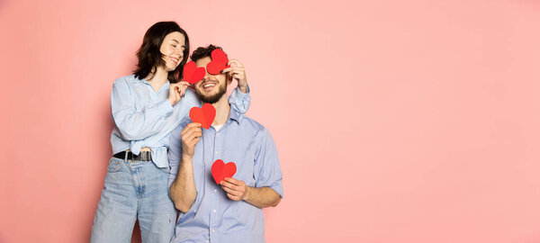 Flyer. Young and happy man and woman holding greeting cards shaped hearts isolated on pink trendy color background. Emotions, youth, love and lifestyle concept