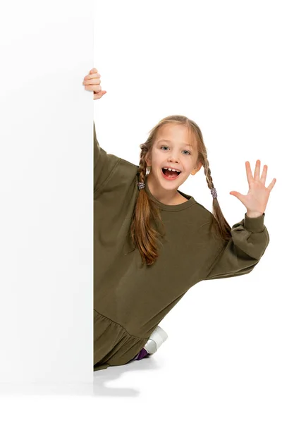 Забавна дитина, маленька дівчинка, учень у повсякденному одязі, що виглядає за стіною ізольовано на білому тлі. Концепція дитинства, емоцій, навчання — стокове фото