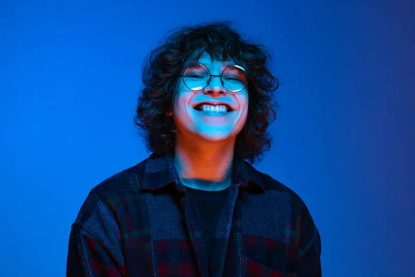 Retrato do jovem homem sorridente, estudante de capuz preto isolado em fundo azul escuro em luz de néon. Conceito de emoções, expressão facial, juventude, aspiração, vendas — Fotografia de Stock