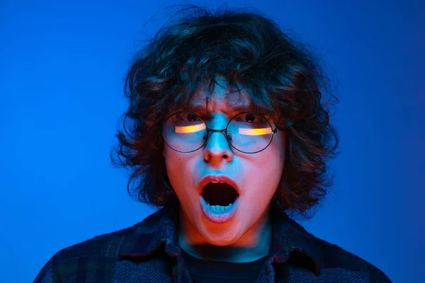 Jovem chocado, estudante de boca aberta olhando para câmera isolada em fundo azul em néon. Conceito de emoções, estudo, educação, moda — Fotografia de Stock