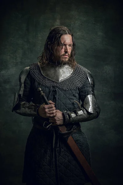 Halvlångt porträtt av brutala seriuos man i bild av medeltida krigare eller riddare med smutsiga sårade ansikte håller stort svärd isolerad över mörk bakgrund. — Stockfoto