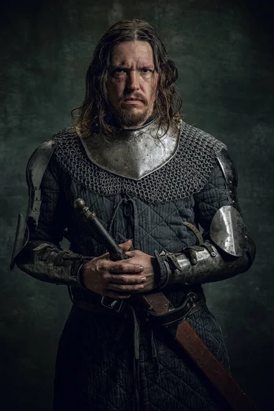 Půl délky portrét brutální seriuos muž v obraze středověkého válečníka nebo rytíře se špinavou zraněnou tváří drží velký meč izolovaný přes tmavé pozadí. — Stock fotografie
