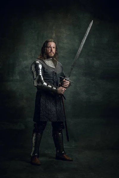 Vintage stijl portret van brutale seriuos man, middeleeuwse krijger of ridder met vuil gewond gezicht met groot zwaard geïsoleerd over donkere achtergrond. Vergelijking van tijdperken — Stockfoto