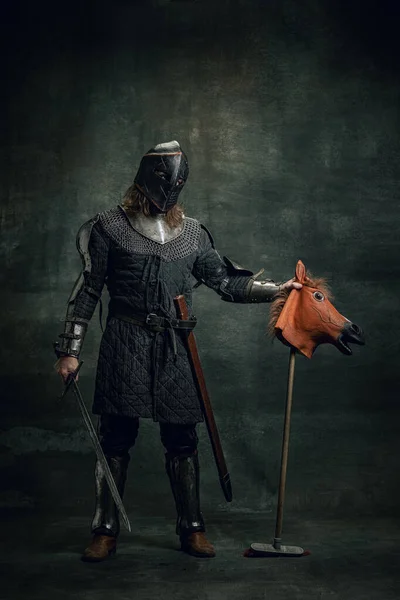 Портрет середньовічного воїна або лицаря в шоломі та обладунках, що їздять на іграшковому коні, тримаючи великий меч ізольовано на темному тлі. Порівняння епох, історія — стокове фото