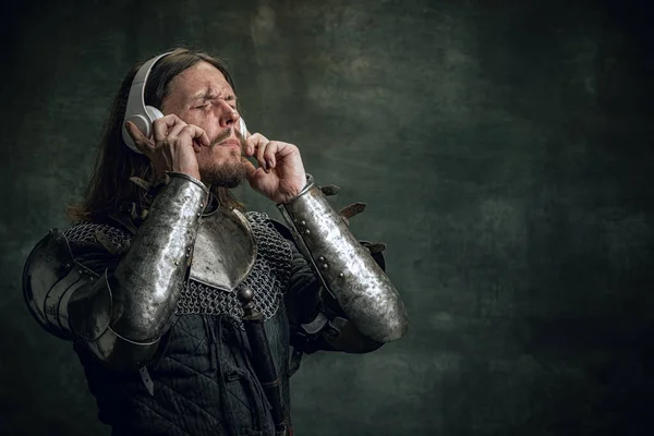 Jovem guerreiro medieval ou cavaleiro com rosto sujo ferido em fones de ouvido ouvindo música isolada sobre fundo escuro vintage. Comparação de eras, história — Fotografia de Stock
