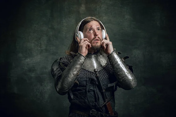 Šokovaný středověký válečník nebo rytíř se špinavou zraněnou tváří ve sluchátkách naslouchající hudbě izolované přes tmavé vinobraní pozadí. Porovnání období, historie — Stock fotografie