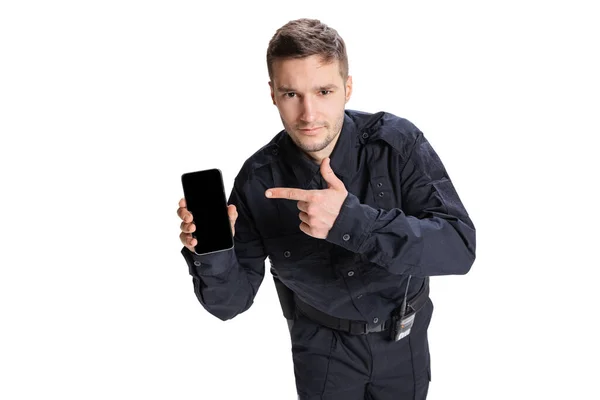 Porträtt av ung man, polis officer klädd i svart uniform med telefon isolerad på vit bakgrund. Begreppet arbete, karriär, lag och ordning. — Stockfoto