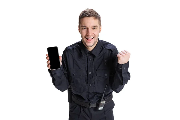 Porträtt av ung man, polis officer klädd i svart uniform med telefon isolerad på vit bakgrund. Begreppet arbete, karriär, lag och ordning. — Stockfoto