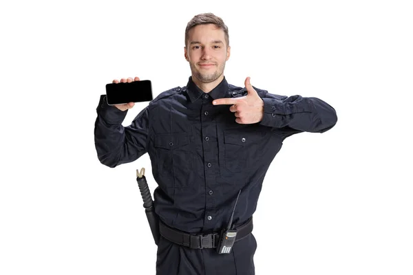 Retrato de homem jovem, policial vestindo uniforme preto usando telefone isolado em fundo branco. Conceito de trabalho, cuidado, lei e ordem. — Fotografia de Stock