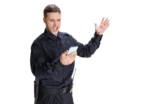 Portrait of young excited man, policeman officer wearing black uniform using phone isolated on white background. Concept of job, caree, law and order. — Stock Photo, Image