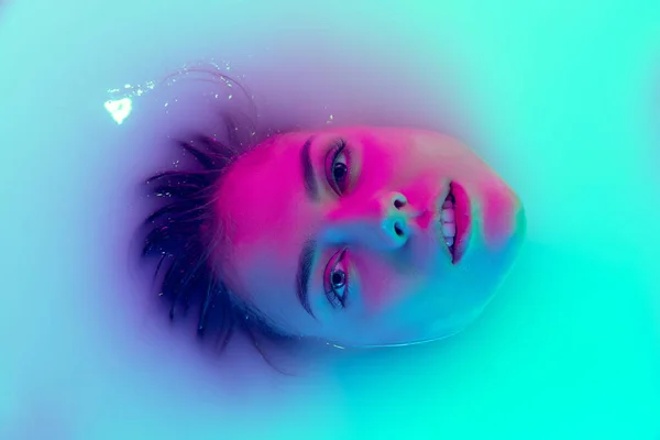 Top view of female face, young beautiful girl in milk bath with soft glowing in blue-green neon light. Beauty, fashion, style, skincare concept — Stock Photo, Image