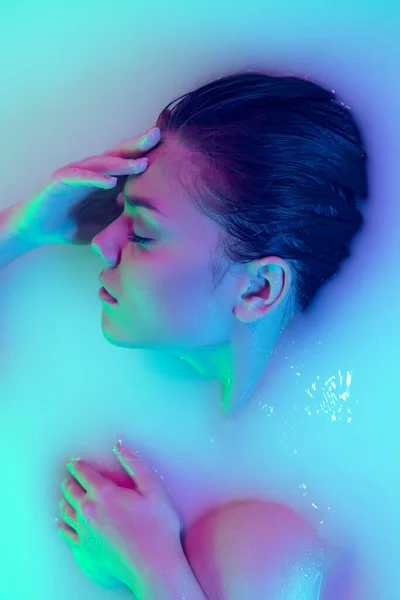 Cara femenina de cerca, joven hermosa chica en baño de leche con suave resplandor en luz de neón azul-verde. Belleza, moda, estilo, concepto de cuidado de la piel —  Fotos de Stock