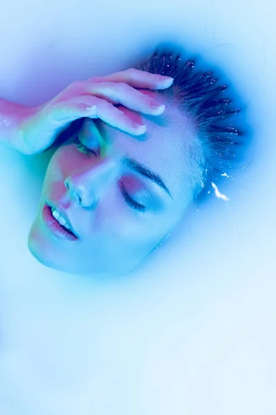 Вид сверху на женское лицо, молодая красивая девушка в молочной ванне с мягкой перчаткой в сине-зеленом неоновом свете. Красота, мода, стиль, концепция Skincare — стоковое фото