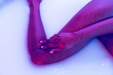 Koyu tenli bir kadın vücudu, süt banyosunda iç çamaşırlı bacaklar ve neon ışıkta yumuşak beyaz bir parıltı. Doğal güzellik, moda, tarz, vücut pozitifliği.