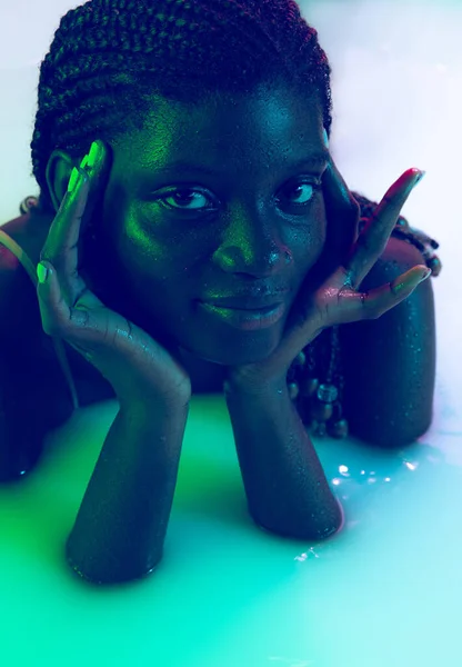 Close-up portrait of beautiful young dark skinned girl lying in milk bath with soft glow in blue-pink neon light. Natural beauty, fashion, style, skincare concept — Stock Photo, Image