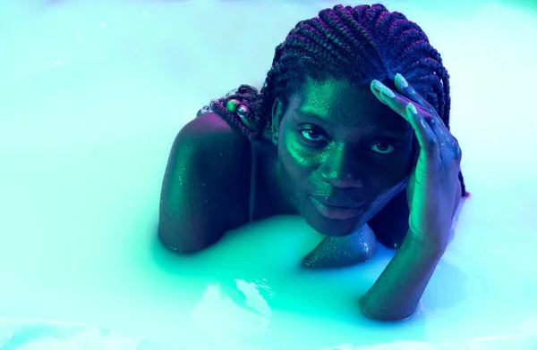 Portrait of beautiful young dark skinned girl lying in milk bath with soft glow in blue-pink neon light. Natural beauty, fashion, style, skincare concept — Stock Photo, Image