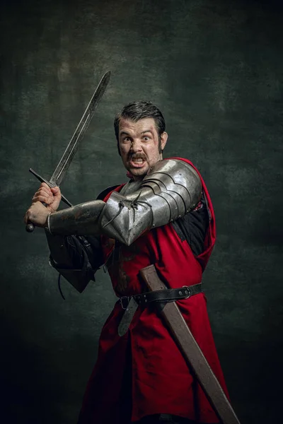 Retrato dinâmico de seriuos homem, guerreiro medieval ou cavaleiro com rosto sujo ferido segurando espada isolada sobre fundo escuro. Comparação de eras — Fotografia de Stock
