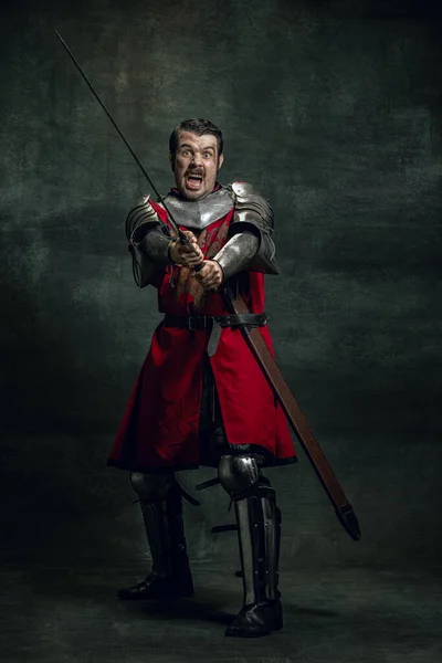 Retrato dinâmico de seriuos homem, guerreiro medieval ou cavaleiro com rosto sujo ferido segurando espada isolada sobre fundo escuro. Comparação de eras — Fotografia de Stock