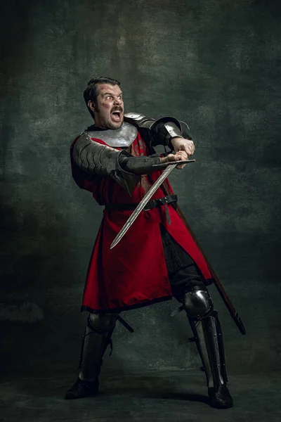 Dynamický portrét seriózního muže, středověkého válečníka nebo rytíře se špinavou zraněnou tváří držícího meč izolovaný na tmavém pozadí. Porovnání období — Stock fotografie